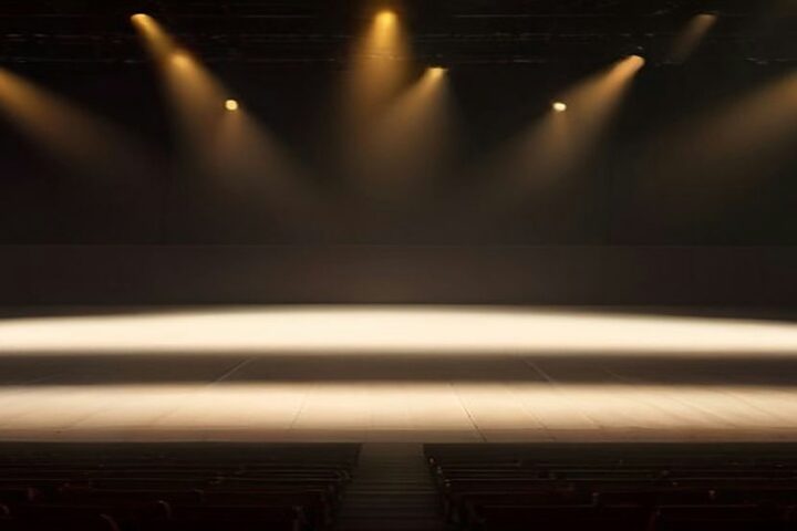 Escenario de concierto vacío con haces de luz ámbar que atraviesan la bruma y filas de butacas sin público en primer plano